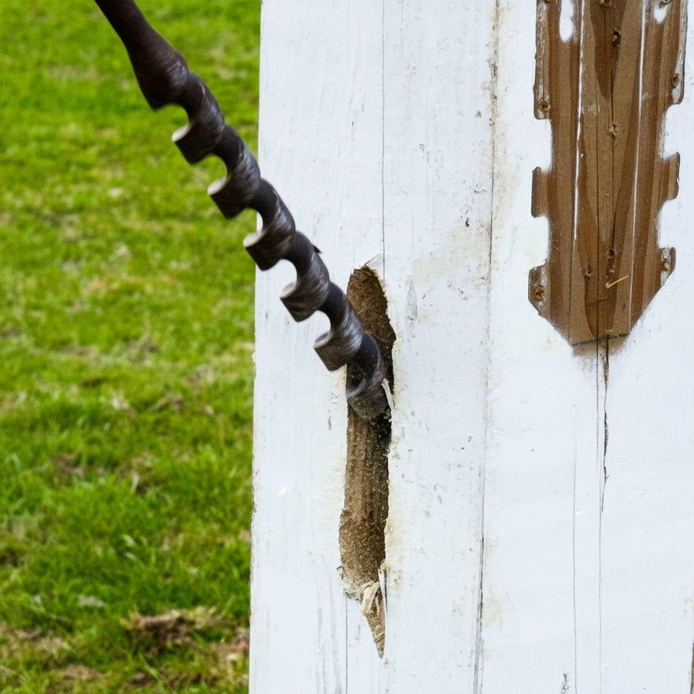 A close-up of a white wooden fence post with a large vertical hole being drilled or channeled into it, with a yellow and black cordless drill partially visible on the left. A matching metal fence panel is attached to the right side of the post, and grass is visible in the background.