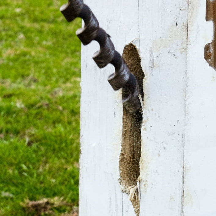 A close-up of a white wooden fence post with a large vertical hole being drilled or channeled into it, with a yellow and black cordless drill partially visible on the left. A matching metal fence panel is attached to the right side of the post, and grass is visible in the background.