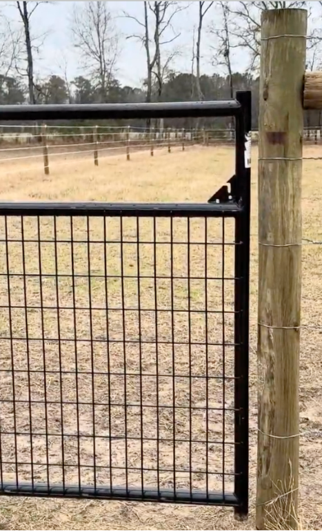 A black metal mesh gate next to a wooden post in an outdoor fenced enclosure with dry grass and trees in the background.